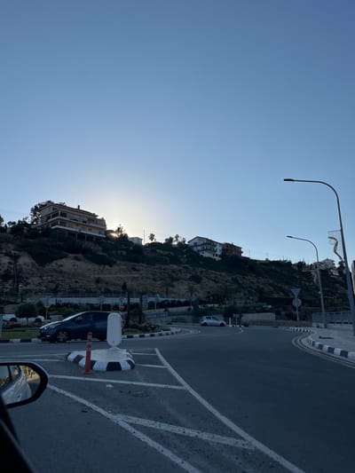 A slightly angled, outdoor shot depicts a road scene near a hillside in Mouttagiaka Community, Cyprus. The sky is a clear, pale blue. A hill, covered with vegetation and scattered buildings, rises from the left side of the image. The sun peeks through the foliage, creating a slight glare.
Two streetlights, with curved arms, are visible along the top edge. A section of road is in the foreground, with painted parking spaces. A dark blue car is parked near a black-and-white striped bollard and an orange traffic cone. The side mirror of the photographer's car is visible in the bottom right corner.