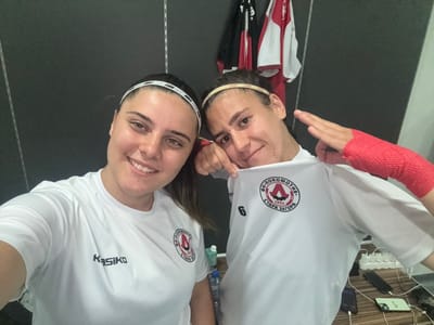 Two young women are posing for a selfie in what appears to be a locker room. Both are wearing white t-shirts with a circular logo on the left chest. The woman on the left has a white headband with black writing. The woman on the right also wears a headband, this one tan with white writing, and has her right hand wrapped in red medical tape. Behind them, several garments, including red and white tops, hang on hooks against a dark gray wall. A shelf or counter runs along the bottom right of the image, holding a power strip with several plugs, two smartphones, and other miscellaneous items. The overall lighting suggests an indoor environment, possibly at a sports facility. There is no visible indication of the specific location within Razgrad, Bulgaria.