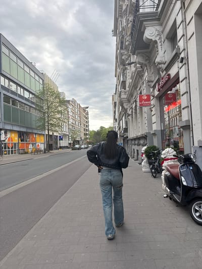 A photograph shows a person walking on a sidewalk in Antwerp, Belgium. The individual is wearing a black leather jacket and blue jeans. A black Vespa-style scooter is parked on the sidewalk nearby. Buildings line the street, with "SIMAS massage" signs visible on one. The sky is overcast. The photo is taken from a low angle.