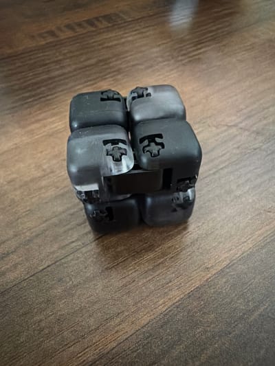 A cluster of black, cube-shaped fidget toys is arranged on a wooden surface. The toys appear to be interlocking, with visible cross-shaped openings on each cube. The wooden surface has a wood-grain pattern with visible lines and variations in color. The lighting suggests an indoor environment. The image was taken in Yablonovskiy, Russia.