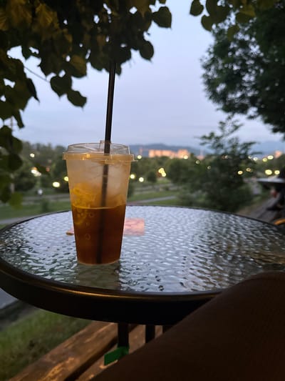 An iced beverage in a clear plastic cup with a black straw sits on a round glass-topped table. The cup contains a dark amber liquid with ice and some yellow fruit slices. The table is set outdoors, with a blurred background of trees, lights, and buildings. The sky is overcast, suggesting it is evening or a cloudy day. The image is taken from a slightly elevated angle, looking down at the table and the drink. Part of a person's leg is visible in the lower right corner of the frame, suggesting they are seated at the table. The location is the city of Vladikavkaz, Russia.