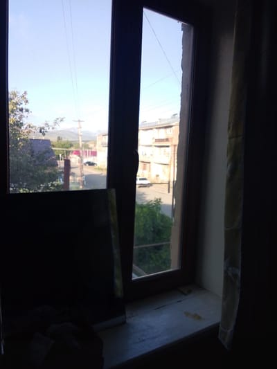 The image shows a view from a window, likely in Sisian, Armenia, looking out onto a street and buildings. The window frame is dark wood, with a partially visible curtain on the right side. Through the window, a clear blue sky is visible, with some power lines stretching across it.

On the left side of the frame, a dark, reflective object, possibly a mirror or glass surface, is placed on the windowsill. It obscures a portion of the view.

The outdoor scene depicts a street with some trees and buildings. A dark-colored car is parked on the street. In the background, there are residential buildings, some with balconies. Mountains are visible in the far distance, suggesting a mountainous region. The overall lighting suggests it is daytime, possibly morning or afternoon. The scene appears to be residential and somewhat rural or suburban.