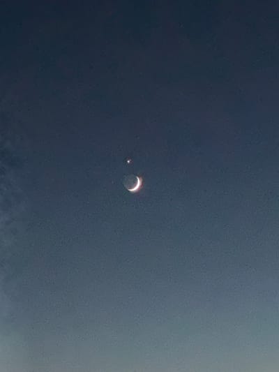 The image shows a very dark, grainy sky with a crescent moon and a bright star visible in the upper center. The moon is illuminated on its left side, showing a thin sliver of light with a pinkish-white glow. Above and slightly to the left of the moon, a small, bright, star-like object is visible. The rest of the sky is a deep, uniform blue-grey, suggesting it is either very early morning or late evening. There are no other discernible objects, people, or structures in the frame, and no indication of the city of Le Brassus, Switzerland.