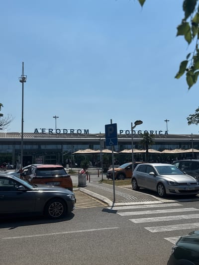 This media file shows the exterior of an airport terminal on a sunny day. The building has large glass windows and a curved roof. Above the entrance, the words "AERODROM PODGORICA" are displayed in large blue letters. In front of the building, several cars are parked, including a silver Volkswagen Golf and an orange car. A pedestrian crossing is visible on the right side of the image, with its white stripes casting long shadows on the asphalt. A blue sign indicating parking and a disabled parking symbol is also present. The sky is clear and blue, suggesting good weather. The overall scene depicts a typical day at an airport, with a focus on the building's signage and the surrounding infrastructure. The time of day appears to be midday, given the strong sunlight and prominent shadows. The location is identified as Golubovci, Montenegro, implied by the signage. There are no discernible people or explicit emotions visible in the media.