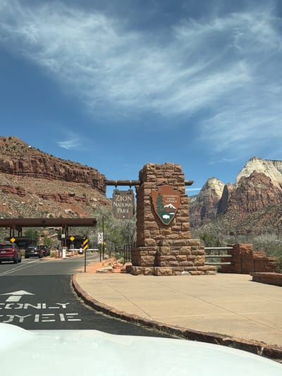 A wide-angle, eye-level shot captures the entrance to Zion National Park on a clear day, framed by towering red rock formations under a bright blue sky with wispy white clouds. In the foreground, a prominent stone monument built from reddish-brown blocks marks the park entrance. Embedded in the stone is the green and brown arrowhead emblem of the National Park Service, while a wooden sign hangs beneath a stone archway, clearly displaying "ZION NATIONAL PARK" in white text.
The scene depicts an active park entry point with multiple lanes. Several vehicles, including dark-colored cars and a red SUV, are queued at entrance booths in the mid-ground. The asphalt road in the foreground features white painted markings, including an arrow and the text "LEFT LANE," indicating the flow of traffic. To the right, part of a white vehicle (likely the observer's car) is visible.
Beyond the entrance infrastructure, the impressive natural landscape of Zion dominates, with steep, stratified red and white cliffs rising dramatically. Sparse vegetation can be seen clinging to the rocky slopes. Various road signs are visible near the booths, including yellow diamond-shaped warnings and red circular signs indicating "NO PARKING" and "NO U-TURN." The bright sunlight suggests it is daytime, likely morning or early afternoon, contributing to a vibrant and inviting atmosphere for visitors entering the park in Springdale, United States of America.
