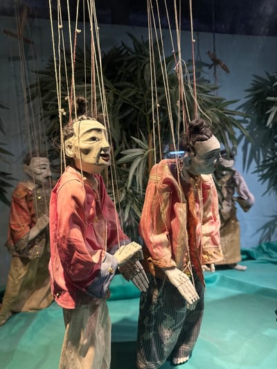 The image depicts several traditional marionettes suspended by strings against a backdrop of dark blue and green foliage. The marionettes appear to be part of a performance or exhibit. In the foreground, two marionettes are prominently displayed. The one on the left has a pale face with distinct facial features, dark hair tied in a bun, and is wearing a pink silk garment with blue cuffs and lighter colored pants. The marionette to its right also has a pale face and dark hair in a bun, dressed in a patterned pink and gold silk jacket over striped pants. Behind these two, another marionette, dressed in a reddish-orange garment, is partially visible, along with a fourth figure in the background, wearing a muted brown and gold outfit. The overall setting suggests an indoor display, possibly a museum or theater, with theatrical lighting. The marionettes are crafted with articulated wooden limbs and detailed facial expressions, evoking a sense of historical or cultural significance.