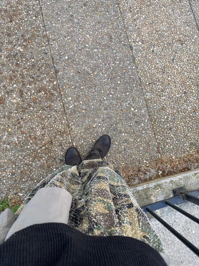 The image shows a top-down view of a person standing on a textured, pebble-like concrete surface. The person is wearing dark shoes and camouflage patterned pants, with a light-colored garment and a dark jacket partially visible. A dark metal railing is situated on a concrete ledge to the right. The location is identified as Les Ulis, France, based on additional (not shown) information.