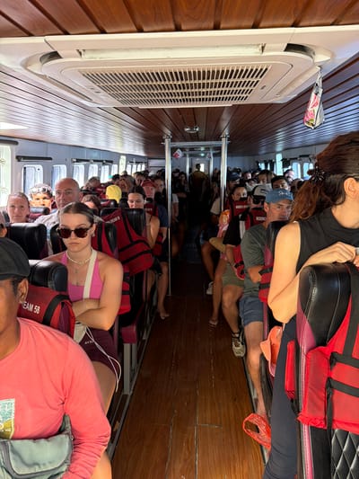 The image shows the interior of a ferry boat, with passengers seated on both sides of a central aisle. The boat has wooden paneling on the ceiling and walls, and an air conditioning unit is visible overhead. Several passengers are wearing red life vests. In the foreground, a woman with sunglasses and a pink top is seated, arms crossed. To her left, a man in a pink long-sleeved shirt and a grey bag is partially visible. On the right, the back of a woman with dark hair is seen. The scene suggests a common mode of transportation, likely within or near a coastal city like Kampung Toyapakeh, Indonesia, given the presence of the ferry and life vests, common for island hopping or inter-island travel in the region.