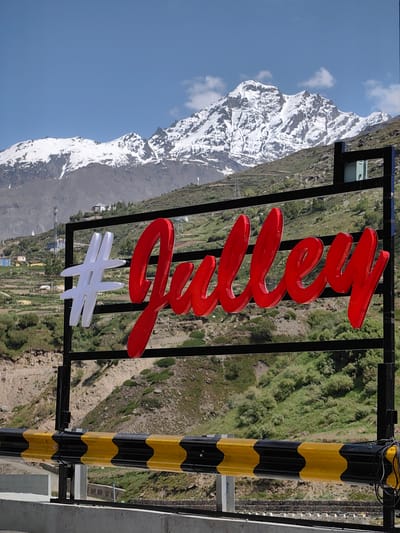 The image depicts a scenic mountainous landscape in Burwa, India, under a clear blue sky with scattered clouds. In the foreground, a prominent black metal frame supports a large, bright red sign with white lettering that reads "#Julle". The sign is illuminated and appears to be made of raised, glossy material. Behind the sign, the mountainous terrain is covered in green vegetation and patches of snow on the higher peaks. Small buildings and structures can be seen scattered on the hillsides. In the immediate foreground, a yellow and black striped barrier is visible, suggesting a road or checkpoint. The overall scene conveys a sense of travel and destination, with the sign acting as a welcoming marker in a beautiful, remote location.