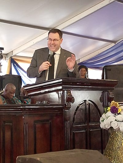 This image captures a lively scene of a speaker addressing an audience under a large white tent, likely in Dushepe, Nigeria. The central figure is a Caucasian man, appearing to be in his 40s or 50s, wearing glasses, a dark suit, a white shirt, and a dark tie. He stands behind a substantial, dark wooden podium adorned with intricate carvings, holding a black microphone in his left hand. His right hand is raised in a gesture, and he has a broad, enthusiastic smile, suggesting he is actively engaging with his listeners.
To the speaker's left, partially visible, is an older man with lighter skin and short, possibly graying hair, wearing a vibrant, patterned garment that could be traditional local attire. He appears to be looking downwards. Behind the main speaker, to his right, a woman with dark skin and a dark head covering is visible, looking forward with a serene expression.
The setting indicates an event of some significance, possibly a community gathering, religious service, or public address. The white tent canopy forms the roof, and decorative blue and gold fabric drapes are visible in the background, adding a festive or formal touch. Large black speakers or other equipment are placed behind the podium on both sides. A floral arrangement featuring white, yellow, and purple flowers in a gold-patterned vase sits to the right of the podium, further enhancing the formal ambiance. The bright, even lighting inside the tent suggests it is daytime or that the area is well-illuminated. No text is visible in the image.