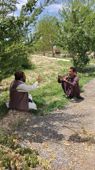 In Qilla Saifullah, Pakistan, two men are in a rural area with trees and vegetation. One man, dressed in a maroon robe, is squatting and looking at a cell phone in his hands. The other man, wearing a brown vest over a white shirt, is kneeling with one arm raised, possibly interacting with the first man. The ground is a mix of grass, dirt, and gravel. A motorcycle is visible in the background near a tree.