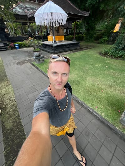 A blonde man with sunglasses perched on his head and a beaded necklace around his neck is taking a selfie in a paved outdoor area. He is wearing a dark grey t-shirt, a yellow sarong tied around his waist, black shorts, and black flip-flops. The man is standing on a paved walkway lined with a grassy area and manicured bushes. In the background, there is a traditional Indonesian temple structure with a large white umbrella adorned with white fringe. The temple features dark wood, a tiled roof, and a raised platform with yellow fabric. The environment appears lush and green, with trees and foliage visible. The scene suggests the man is visiting a cultural site in Dawan, Indonesia.