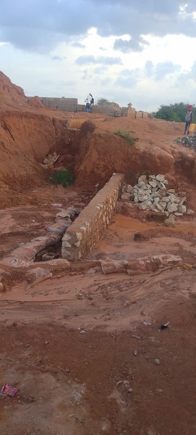In Rayfield, Nigeria, an overcast sky looms over a construction site with red earth and piles of rocks. Two individuals are visible in the distance, near unfinished concrete structures. In the foreground, a partially constructed stone wall runs across the scene, with debris and muddy ground surrounding it. Another person, carrying a yellow bucket, walks away from the camera towards the right side of the image.