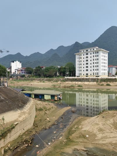 The image depicts a low river scene in Thành phố Hà Giang, Vietnam. In the foreground, a concrete retaining wall borders a channel with stagnant water and debris. Adjacent to the channel is a small floating structure. The river itself reflects a large, white building located on the opposite bank. This building is a multi-story structure with rows of windows. In the background, a range of tree-covered mountains rises behind the town. The sky is overcast.