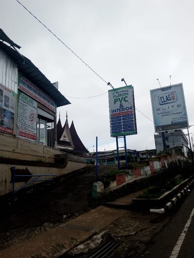 The scene depicts a street in Thotagamuwa, Sri Lanka, under an overcast sky, viewed from a slightly elevated position looking up an incline.
On the left, a multi-story building with a corrugated metal upper facade features several red and white banners. A prominent banner displays "GEMILANG PLAFON PVC & EKSTERIOR" and contact information. Below it, another sign lists "GEMILANG PLAFON PVC" services. Below this building, a concrete retaining wall supports an earth bank. A blue metal arrow sign points to the right. A concrete access ramp or pathway, featuring a metal drainage grate in the foreground, leads up the slope. Behind the initial building, a structure with a distinctive, multi-tiered, steeply-pitched dark roof, characteristic of traditional Indonesian architecture, is visible.
On the right, an asphalt road with a white solid line on its left edge runs parallel to the incline. A concrete curb separates the road from a planter strip with sparse vegetation, partially painted in sections of red, white, and green/blue. Two large billboards stand prominently on tall poles. The closer billboard, framed in green, reads "GEMILANG PLAFON PVC & INTERIOR" and lists various interior services such as "PLAFON HOLO," "PARTISI PVC," "KUSEN PVC," "LANTAI VINYL," "WALLPAPER," "GAZEBO," and "PINTU PVC." The second billboard, framed in white, advertises "NEW CLAS MILD ELITE INTENSE SMOOTHNESS" and includes the Indonesian health warning "Merokok Membunuhmu" (Smoking Kills You). In the background, beyond the billboards, multiple urban buildings are visible. Overhead, power lines or communication cables traverse the frame. No individuals are present in the scene. The language on all visible commercial signs is Indonesian.