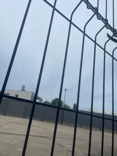 The image shows a view through a black metal fence. The fence bars are positioned diagonally across the frame, with some horizontal elements at the top. In the background, there is a grey corrugated fence or wall, and beyond that, a low-rise building with a white facade and a darker roof. Trees are visible to the right of the building. The sky is overcast and appears light blue-grey. The ground in front of the corrugated fence is paved with concrete, showing some grid lines. The scene appears to be an outdoor area, possibly a yard or parking lot, likely in an industrial or commercial setting. The city of Kołobrzeg, Poland, is not explicitly visible or indicated within the image itself.
