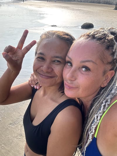 Two women are posing for a selfie on a beach. One woman has braided hair and is wearing a swimsuit with neon green straps. The other woman has dark hair and is wearing a black swimsuit. The woman in the black swimsuit is holding up a peace sign. They are standing on a sandy beach with wet sand and some rocks visible in the background. The location is Jimbaran, Indonesia.