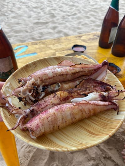 A close-up, eye-level shot shows a plate of grilled squid on a beachside table, likely in Cam Lam, Vietnam. The squid, lightly grilled with some char marks, are arranged on a tan, wood-patterned plate. Two small dipping dishes, one with a pink sauce and the other with a darker sauce, are visible on the table behind the plate. In the background, two brown beer bottles with green caps stand upright, and the sandy beach and ocean can be seen. The scene suggests a casual dining experience outdoors.