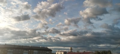 The image captures a dramatic sky over a town, likely Volchansk, Russia. The scene is dominated by a vast expanse of clouds. The clouds are a mixture of light and dark grays, with some patches of bright white where the sunlight is catching them. They are textured and voluminous, suggesting a dynamic weather system. Some thin, wispy clouds are scattered across the lighter blue sky, while thicker, more ominous clouds gather towards the horizon.
Beneath the sky, the rooftops of several buildings are visible. On the left, a long, multi-story apartment building with a dark roof is prominent. Its facade appears to be light-colored, with visible windows and balconies. Further to the right, a building with a golden dome and a cross on top stands out, hinting at a church or religious building. Another building with a red roof is also visible. The presence of these structures suggests a populated area, likely a town or city.
The lighting indicates that it is either early morning or late afternoon, possibly around sunset, given the warm glow on some of the clouds and the contrast between the illuminated and shadowed parts of the sky. The overall mood of the image is one of natural grandeur and perhaps a touch of drama due to the turbulent cloud formations. There is no visible text in the image.