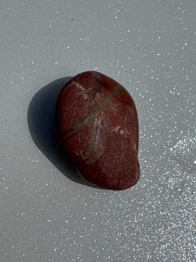 This media file displays a single, oval-shaped, reddish-brown stone resting on a surface covered in fine, reflective glitter. The stone has a rough texture and appears to have lighter-colored veins or inclusions running through it. The glittered surface is a light grey color, and the glitter particles create a shimmering effect under bright lighting. The image is a close-up shot, with the stone positioned slightly off-center, casting a subtle shadow to its right. The lighting appears to be strong, possibly sunlight, as indicated by the bright reflections on the glitter and the distinct shadow. There is no visible text in the image. The scene is static and focuses entirely on the texture and appearance of the stone and its sparkly background. The location context provided, Добровское сельское поселение, Ukraine, does not offer any visual cues within the image itself.
