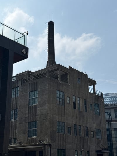 The image depicts a vertical, low-angle view of architectural structures against a blue sky with some cloud cover. A tall, gray concrete building with multiple windows and a smokestack protruding from its side dominates the scene. The building exhibits a somewhat dilapidated or aged appearance. In the lower portion of the image, a modern building with a black facade and the number "4" prominently displayed on it is visible. A security camera is mounted on the modern building's exterior. The structure in the upper right of the frame has lettering reading "ZE GALLERY". The location is Hongkou District, China. No people are visible in the image.