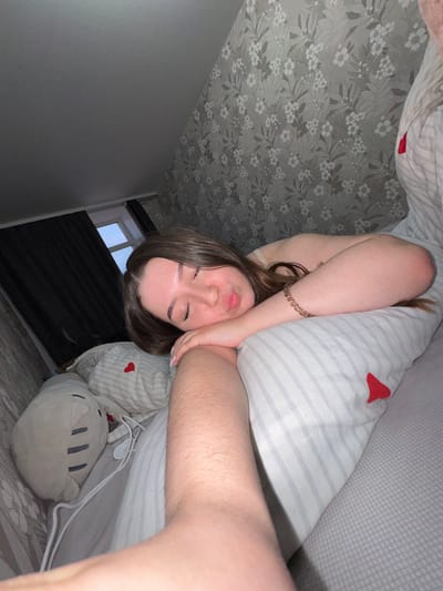 An indoor image captures a young adult female lying on a bed in a bedroom located in Osentsy, Russia. She has long brown hair and fair skin, with her eyes closed and lips slightly parted, appearing to be asleep or resting. She wears a light-colored camisole or tank top. Her right arm, adorned with a multi-beaded bracelet, is bent with her hand resting near her head on a pillow. Her left forearm extends towards the camera, indicating a close-up or self-portrait perspective.

The pillow beneath her head is white with light blue horizontal stripes and features a small red heart appliqué. Another partially visible pillow also has a red heart. The bed is covered by a light grey sheet with a subtle grid pattern. To her left, a light grey plush toy resembling a "Pusheen" cat is visible, along with white charging cables.

The wall behind her is covered with light-toned floral wallpaper featuring white and darker botanical patterns. The ceiling above is plain and light grey, appearing to slope. In the background, a window with a white frame is partially obscured by dark curtains, revealing a soft blue sky outside, suggesting either twilight or early morning light.