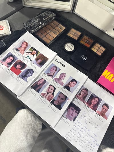 The image displays a flat lay of items on a dark surface, likely a table covered with a black towel, indicating a professional workspace, potentially a backstage or studio setup in Issy-les-Moulineaux, France, though the exact address is not discernible. The primary subjects are several sheets of paper, acting as model casting or reference sheets, laid out horizontally.

These sheets feature headshots and half-body portraits of individuals, predominantly women, with names (e.g., Jeanne, Roxane, Noa, Angelique, Daniela, Mathias, Martin) and measurements (height, chest, waist, hips) printed below each image. Many entries have handwritten checkmarks or notes, such as "look," "bleu," and "violet," suggesting selection or specific styling considerations. A date "19/01/2023" is handwritten on one sheet. Extensive handwritten notes in French are present on the bottom right sheet, detailing makeup instructions or model observations, including mentions of "Pour le lineup," "MAC creme pour le corps," "Daniela Allergique crene," and descriptions of "deux looks."

Various makeup products are arranged on the black towel in the upper right quadrant. These include multiple eyeshadow or foundation palettes, some appearing used, a round container labeled "NYX HD Finishing Powder," and a black rectangular palette case marked "NUMERIC PROOF." A clear plastic case filled with makeup brushes is positioned next to a large palette. A white packet of "Lingettes démaquillantes" (makeup remover wipes) is visible on the far left. A portion of a bright pink item, possibly a folder or book with "MATIS" visible, is on the far right. A white towel is draped over a chair or object in the lower-left foreground. The lighting is even, suggesting an indoor, controlled environment.