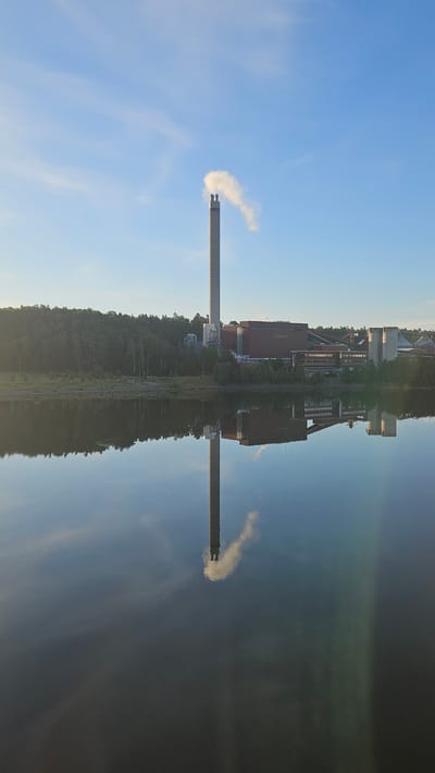 This is a vertical, eye-level, full shot of a power plant, likely a cogeneration plant, on the shore of a body of water. The plant features a tall, cylindrical, gray smokestack emitting a plume of white steam against a clear blue sky with some wispy clouds. To the right of the smokestack is a large, red, rectangular building with the text "Södertörn" visible, indicating the location is Södertälje, Sweden. Several other industrial structures, including concrete silos and smaller buildings, are situated behind the red building. The plant is surrounded by a dense forest on the left and extends towards the right. The water in the foreground, likely a river or lake, perfectly reflects the smokestack, the steam plume, and the industrial buildings. The reflection creates a symmetrical composition, with the water surface appearing calm and smooth. The overall lighting suggests it is early morning or late afternoon due to the soft light and clear sky. There are no people visible in the image.