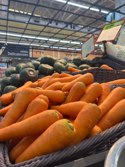 The image shows a close-up view of a basket filled with fresh carrots in what appears to be a grocery store or market. The carrots are bright orange and have a slightly tapered shape, with visible root ends. Behind the carrots, there are more vegetables, including several round, dark green squash or pumpkins, also piled in a display. In the background, overhead lighting fixtures and store shelving are visible, along with a sign that reads "Delicatessen". There are also price tags visible, one of which displays the number "59". The lighting suggests an indoor setting, likely a commercial establishment. The scene depicts a produce section of a store.