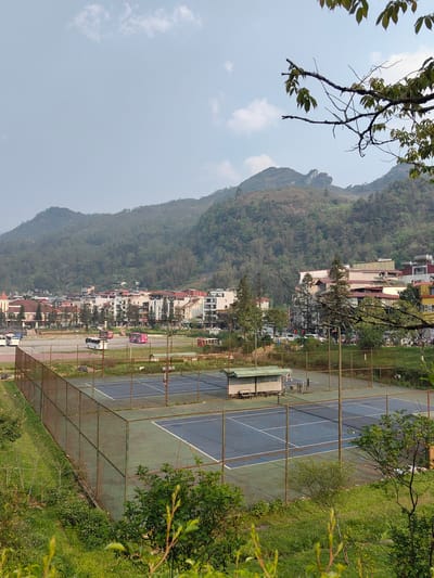 The image presents an elevated view of a recreational and urban area within Sa Pa, Vietnam, set against a mountainous landscape under a partially cloudy sky.
In the immediate foreground, green foliage and tree branches with leaves partially obscure the lower section of the frame. Below this vegetation, a rusted chain-link fence encloses two outdoor tennis courts. The courts feature blue playing surfaces with white lines and green outer boundaries. A small, light-green-roofed shelter with benches is positioned at the far end between the two courts. One indistinct individual is visible on the far court near the baseline, and another fainter figure appears on the near court.
Beyond the tennis courts, a large, open area of sparse grass and dirt accommodates several parked vehicles, including multiple white and pink tour buses, and private cars. Further into the midground, a dense collection of multi-story buildings, characterized by varied architectural styles and colors (including red-roofed structures and modern white facades), forms an urban cluster. A distinct building with a red-roofed turret is visible on the far left of this urban section. Green trees are interspersed among the buildings.
The background is dominated by steep, heavily forested mountains, exhibiting peaks and ridges. A prominent rocky outcrop is discernible on one of the mountain summits towards the left. The sky is a light blue with scattered white clouds. The scene captures a segment of Sa Pa from a vantage point overlooking recreational facilities and the town's development nestled within its mountainous terrain.