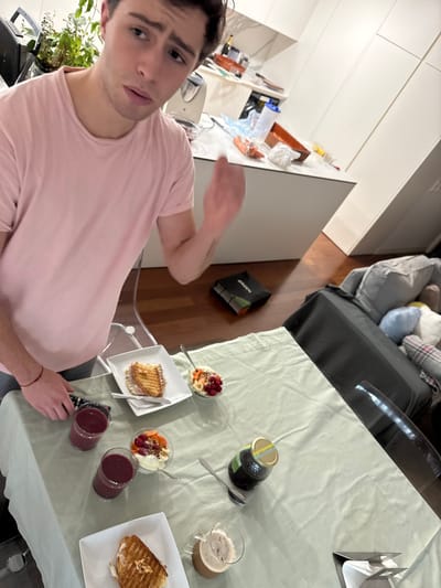 An indoor scene in Nogueira e Silva Escura, Portugal, captures a young Caucasian male standing behind a table set with breakfast items. The male, positioned in the foreground, wears a light pink short-sleeved t-shirt and a dark wristband on his left wrist. He has dark, short hair and a neutral expression, with his mouth slightly ajar. His right hand is raised and partially blurred, indicating motion. His left hand rests near the table.
The table is covered with a light green tablecloth and displays multiple food and drink items. These include two white rectangular plates, each holding a grilled sandwich. Two clear glass bowls contain what appears to be yogurt or oatmeal topped with various fruits (berries, melon, kiwi) and granola. Two clear glasses hold a dark purple-red liquid, likely smoothies or juice. A clear mug contains a light brown, frothy beverage, possibly coffee or a latte. A dark green jar with a yellow lid, presumably containing honey or a similar condiment, rests on the table with a spoon beside it.
In the background, a modern kitchen area is visible with white, minimalist cabinets and countertops. A silver kettle and other kitchen items are present on the counter. The floor is polished wood. To the right of the table, a grey sofa with multiple cushions and throws suggests an adjacent living space. A black bag with "NATRO" or "NATTRO" branding lies on the wooden floor between the table and the kitchen counter.