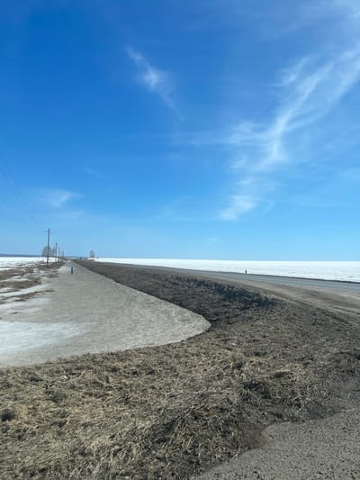 The image shows a rural landscape under a bright blue sky with wispy clouds. A straight, slightly snow-covered road runs through the center of the frame, extending into the distance. One side of the road is covered in snow-dusted terrain, while the other side consists of dark, dry grass and vegetation. Power lines with utility poles are visible along the roadside. The location is not specified as Kueda, Russia.