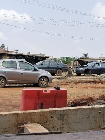 The image depicts an outdoor area in Magboro, Nigeria, characterized by vehicles and structures on an unpaved, reddish-brown dirt surface beneath an overcast sky.
In the immediate foreground, a red plastic road barrier, consistent with a water-filled barrier, is centrally positioned on a concrete base. A red cap is visible on the barrier's top surface, alongside a small, dark, crumpled object.
Behind the barrier, a silver five-door hatchback vehicle, identified as a Toyota Yaris/Vitz model, with mud-splashed wheels, is parked facing right. To its rear-right, a dark blue sedan, consistent with a Peugeot 406, is parked, appearing to be missing its front driver-side wheel and resting directly on the ground. Further to the right, another dark sedan is parked with its hood open, indicating a state of repair or disassembly. This vehicle also appears to be missing its front driver-side wheel, resting on unseen supports or the ground. The midground consists of uneven, reddish-brown earth with visible ruts.
In the background, several single-story structures, likely workshops or storage areas, with corrugated roofs, are visible. These structures are interspersed with sparse green vegetation, including a palm tree. Multiple utility lines stretch horizontally across the pale, cloudy sky. A utility pole with transformers is visible on the far left. No individuals are present in the scene.
