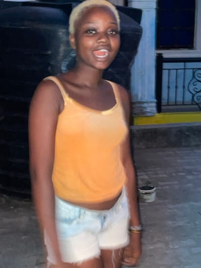 A young woman with platinum blonde hair and a yellow tank top lies on a dark surface. She wears light blue and white shorts, and her mouth is open in a smile or shout. Behind her, there is a building with a window and a wrought-iron design above. The background includes a light-colored wall. The photo has motion blur, indicating movement when the image was taken.