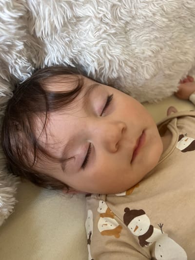 A baby is sleeping on a fluffy, light-colored rug and a light-colored surface, possibly a couch or bed. The baby is wearing a tan-colored top with illustrations of snowmen and is lying on its back with its eyes closed. The baby has dark hair. There are no identifiable landmarks or characteristics to place this image within the city of Elena, Bulgaria. The focus is on the sleeping child.
