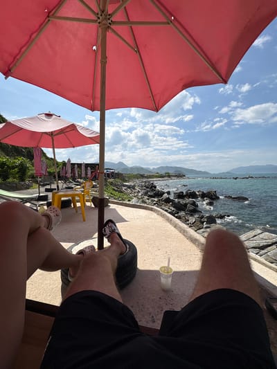 An overhead shot captures two individuals enjoying a seaside view in Vĩnh Lương, Vietnam, seated under a large pink umbrella. One person, wearing pink Crocs, has their legs crossed while resting on a repurposed tire. The other, wearing black and white patterned slides, also has their feet on a tire, with black shorts visible. In the background, a turquoise sea meets a rocky coastline under a partly cloudy sky. A small yellow table and chairs are set up nearby, along with other folded umbrellas and lounge chairs, suggesting a beachside establishment. A cup with a straw sits on the concrete ground next to the seated individuals, hinting at a refreshing drink.