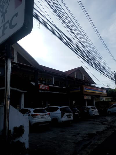 The image depicts a view of a street scene with a KFC restaurant in Phou Khoun, Laos. The building housing the KFC also has an Indomaret store attached to it. Several cars are parked in front of the establishment. Overhead, there is a dense network of power lines. The picture is taken during the day, but the light is dim, creating dark shadows in the foreground. A portion of a KFC sign is visible in the left corner of the frame.