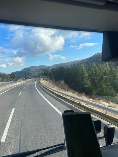 The image provides a first-person view from a vehicle, looking forward through the windshield onto a multi-lane highway near Arbúcies, Spain. The asphalt road surface shows two visible lanes in the direction of travel, delineated by white dashed and solid lines, and curves gradually to the right. A metal guardrail separates the highway from an ascending embankment on the right, which is covered with dry, light-colored grass and a dense stand of dark green coniferous trees. In the background, undulating forested hills extend across the horizon under a bright blue sky dotted with scattered white cumulus clouds. A dark-screened smartphone or tablet, secured in a dashboard mount, is visible in the lower right foreground. A portion of the vehicle's interior trim is visible at the top edge of the frame.