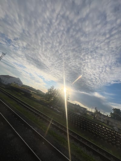 The image depicts a railway track in the city of Kauchuk Stantsiya, Russia. Multiple parallel railway tracks run diagonally from the foreground towards the background. The tracks are laid on gravel with green grass growing between and beside them. On the left side of the tracks, a large white building with a red roof is visible, along with what appears to be a yellow construction vehicle or crane. Further in the background, several multi-story residential buildings with light-colored facades and visible windows are present, interspersed with trees and other vegetation. The sky is filled with a dramatic cloud formation, characterized by a patterned arrangement of small, white clouds against a blue sky. The sun is positioned in the upper right quadrant of the image, casting a strong lens flare and a rainbow-like refraction across the scene, indicating it is likely early morning or late afternoon. No people are visible in the image.