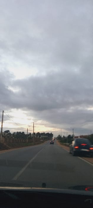 An overcast sky looms over a straight road. Several vehicles, including one with illuminated taillights, are visible. Utility poles line the side of the road where a small strip of green space is followed by sparse trees. The view is likely taken from inside a vehicle, with part of the vehicle visible in the lower right corner of the frame.