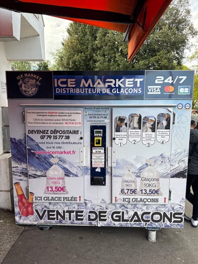 The image shows an "Ice Market" ice vending machine located in an outdoor setting, likely a street. The machine is labeled "Distributeur de Glaçons" and offers ice in bags of 5kg (6.75€) and 10kg (13.50€). The machine also sells 10kg of crushed ice for 13.50€. The vending machine accepts Visa, Mastercard and Carte Bancaire. It indicates it's open 24/7. Contact information is provided, including a phone number and website. The machine has instructions on how to purchase ice. The phrase "Vente de Glaçons" is prominently displayed. The location of the business is Pantin not Les Ulis. People are visible in the background. A red awning and some trees are also in the background.