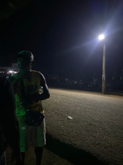 The image depicts a street scene at night in Agba Umana, Nigeria. A person wearing a yellow jersey and light-colored shorts is standing on an asphalt surface. A street light illuminates the area. In the background, there is a gathering of people. A plastic bottle is visible near the person.