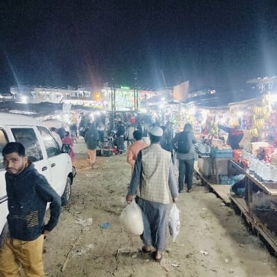 This image depicts a bustling night market scene in the city of Bhola, Bangladesh, specifically near a river port or ferry terminal. The ground is unpaved, dusty, and strewn with litter including plastic bags. Bright artificial lights illuminate numerous makeshift market stalls lining the right side of the path, displaying goods such as bananas, bottled water, and various packaged items. In the background, a large, brightly lit white passenger ferry with multiple decks is visible, likely docked.
Approximately 15-20 individuals are present. In the left foreground, a male with dark hair and a beard, wearing a dark hoodie with yellow stripes and brown pants, stands looking towards the camera, his left hand on his hip. To his right, a white multi-purpose vehicle is parked. In the mid-ground, a male wearing a white skullcap, a light plaid vest over a dark tunic, and dark pants walks away from the camera, carrying a large white bag in his left hand and a smaller white bag in his right. To the far right, partially obscured, a vendor in a red and black top is seen arranging produce, specifically bananas, on a stall. Other individuals are walking and congregating throughout the market area.
