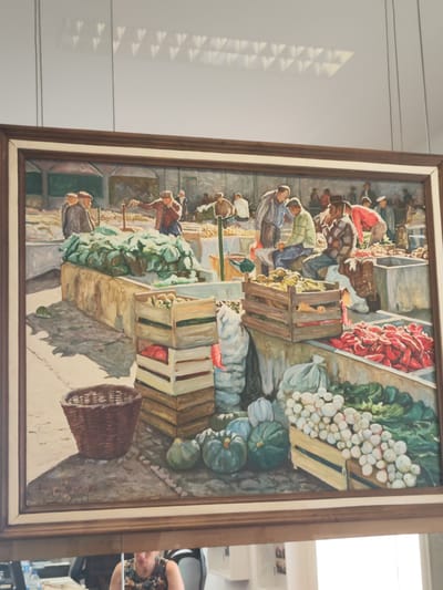 This is a painting depicting a market scene, likely in Ribeira Grande, Portugal. In the foreground, there are crates filled with produce, including large heads of lettuce, red peppers, and white onions or garlic. A woven basket sits to the left, casting a shadow. Several people are shown in the middle ground, some standing and others seated, engaged in what appears to be selling or buying produce. They are dressed in casual, somewhat dated clothing. The background shows more stalls and a hint of an outdoor setting with what could be market structures. The painting style is realistic, with visible brushstrokes and a muted color palette. The reflection in the mirror below the painting shows a person in a floral top.