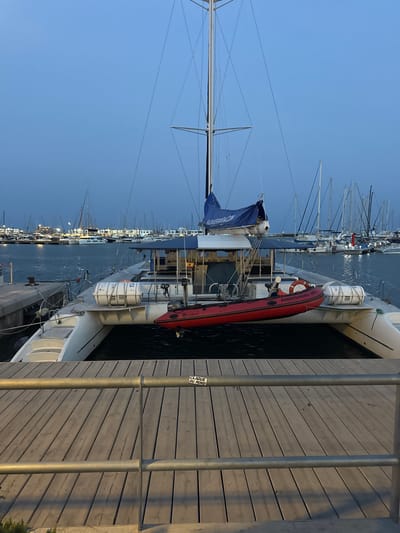 The image depicts a catamaran docked at a harbor in Alicante, Spain, during twilight. The catamaran, a white vessel with multiple hulls, is the central subject. A red inflatable dinghy is secured to its side, and a life raft is visible on the deck. The masts and rigging of the catamaran and other sailboats in the background are silhouetted against the pale blue sky. The foreground shows a wooden boardwalk with metal railings, suggesting a public area. The water is calm, reflecting the faint lights from the boats and the distant cityscape. The overall atmosphere is peaceful, capturing the quiet of a harbor at dusk.