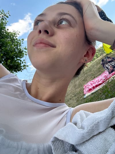 A young woman is shown from a low angle, looking upwards with her hand on her head. She is outdoors, with a tree and blue sky with clouds visible in the background. A pink and white striped towel or blanket is visible on the grass behind her. The woman is wearing a sheer, light-colored top and a towel is draped over her shoulder. The image appears to be taken in Bürgstadt, Germany.