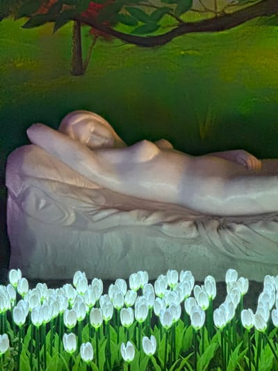 A close-up shot of a statue or art installation. The statue is of a female figure, possibly made of marble or a similar material, positioned against a backdrop of green foliage and a column of white flowers. The location is Binh Lu, Vietnam.