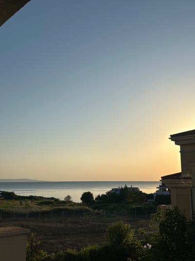 The image depicts a coastal landscape in Chernomorets, Bulgaria, at sunset. A clear, light blue sky transitions to a soft yellow and orange hue near the horizon, indicating the setting sun. In the foreground, a partially visible building with a tiled roof and beige facade is on the right. Below the building, there is dark soil with scattered green bushes and trees. A fence separates this area from a grassy slope covered with wild vegetation. Beyond the slope, the calm sea stretches to the horizon, with a faint landmass visible in the distance. Several modern buildings, possibly hotels or apartment complexes, are nestled among trees on the coastline. The overall scene conveys a tranquil, serene atmosphere of a coastal evening.