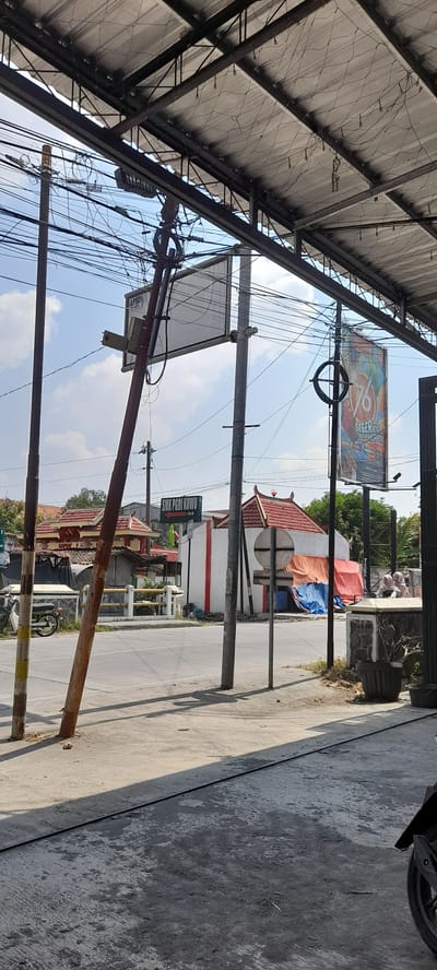 The image depicts a street scene in Crewek, Indonesia, under a bright, partly cloudy sky. A prominent, leaning utility pole with numerous wires dominates the left side of the frame, displaying a sign that reads "SMK PGRI KUWU" with an arrow pointing to the right. To the right of this pole, another utility pole supports a large, rectangular white sign with some text and a smaller rectangular light fixture. Further to the right, a tall, vertical banner with the number "76" and Indonesian text, possibly related to an anniversary or event, hangs from a pole.

In the background, there are several buildings with traditional red tiled roofs. One building, painted white with red trim, appears to be part of the school mentioned on the sign. Tarpaulins in orange and blue are draped over some structures. A few people, possibly students in headscarves, are visible in the far right background near a building and a potted plant. A motorcycle is parked on the far left of the image. The foreground consists of a concrete sidewalk and road, with shadows cast by the overhead structure. The overall impression is a typical street view in a residential or educational area of a town in Indonesia.