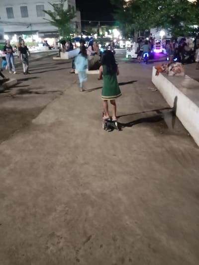 The image shows a young girl riding a scooter on a paved area in front of a building in Kaliwungu, Indonesia, located at -6.962 latitude and 110.253 longitude. The girl is wearing a green dress and her hair is pulled back from her face. The scooter is red and black and has a small, round, black seat. The girl is facing away from the camera and is riding towards the left. The pavement is grey and appears to be slightly wet. There are other people in the background, some of whom are walking and some of whom are sitting on benches. The building in the background is a two-story building with a white facade. There are trees on either side of the paved area and the background is obscured by darkness.